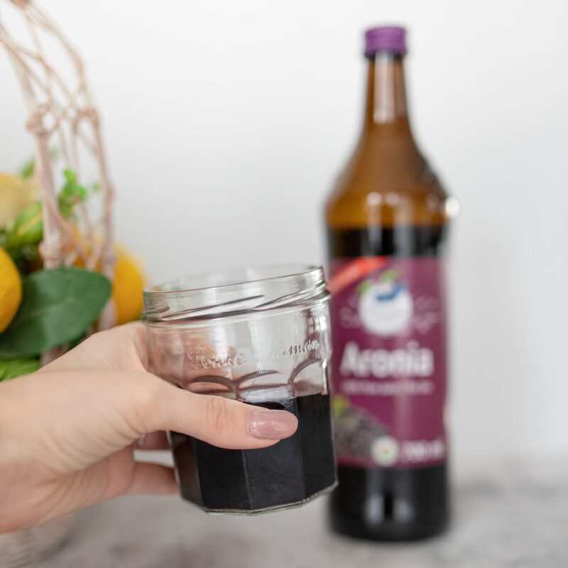 Person holding a glass of Aronia juice in front of an Aronia Original juice bottle
